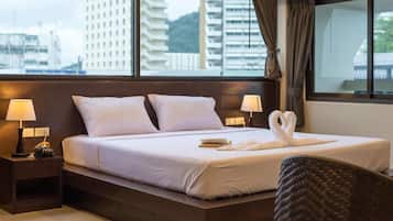 Quarto de hotel com uma cama grande, duas luminárias de cabeceira, vista para prédios pela janela e uma cadeira em primeiro plano.