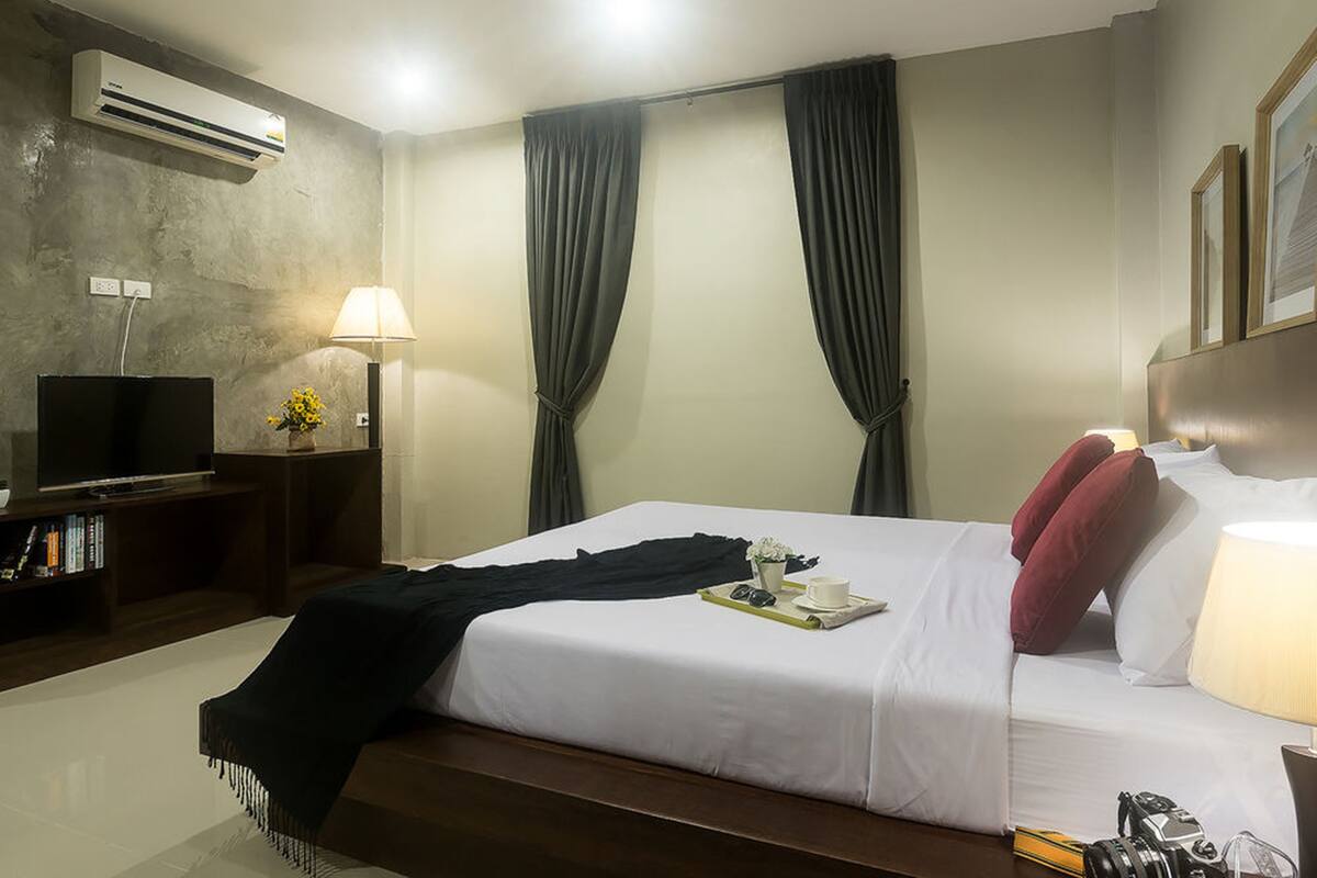 A hotel room with a bed, a television, a desk with a lamp, and an air conditioning unit.