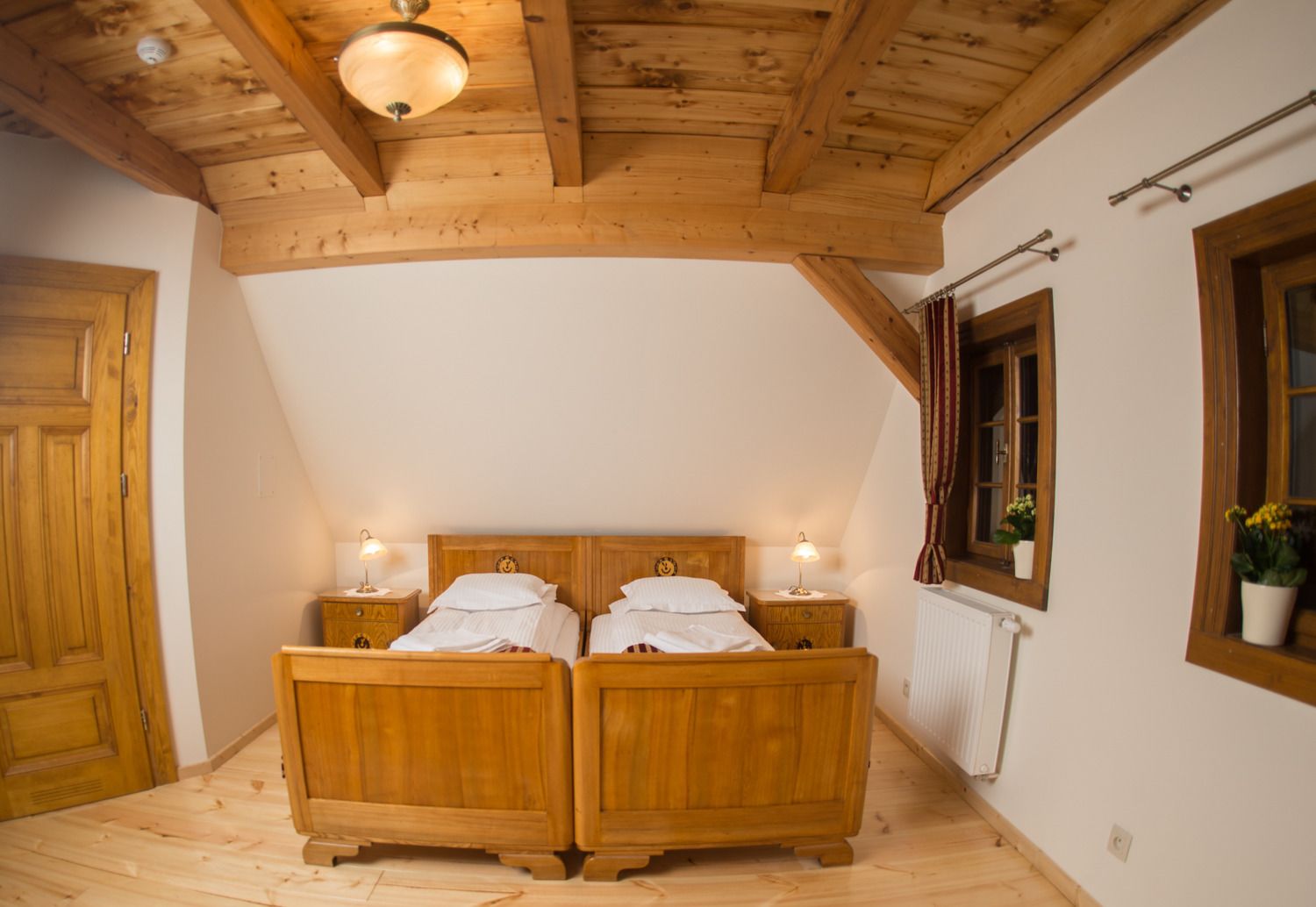 A wooden bedroom with a sloped ceiling, two beds, bedside lamps, a radiator, and a window with curtains.