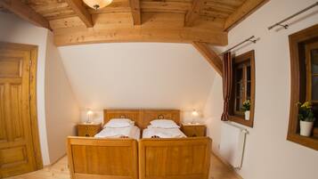 A wooden bedroom with a sloped ceiling, two beds, bedside lamps, a radiator, and a window with curtains.