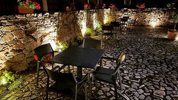 An outdoor patio with stone flooring, metal tables, and chairs, surrounded by stone walls and potted plants.