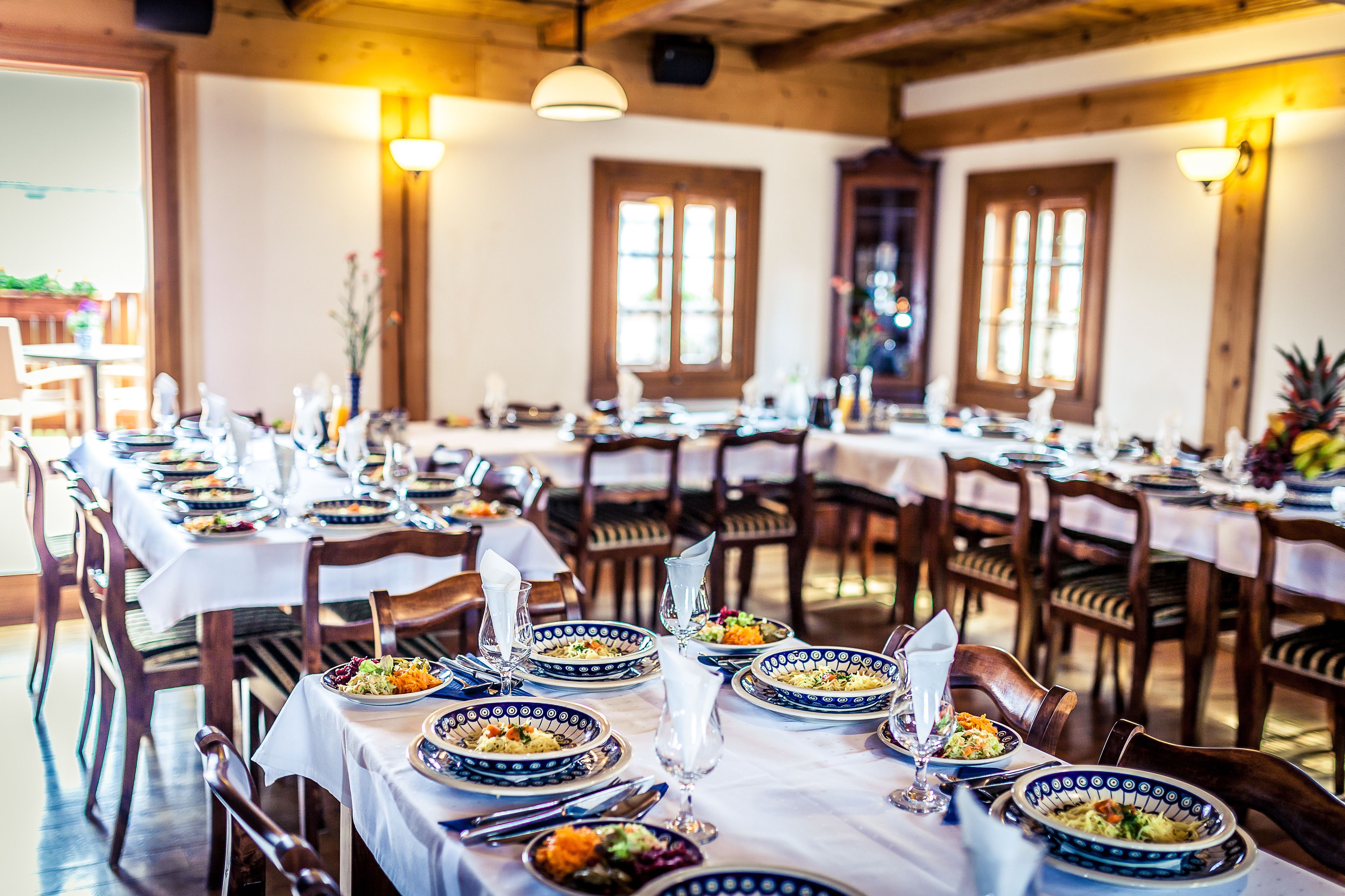 Un espace repas avec plusieurs tables dressées pour les repas, des chaises en bois et de grandes fenêtres laissant entrer la lumière naturelle.