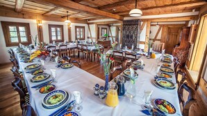 Une grande salle à manger avec de longues tables dressées pour le repas, des poutres en bois au plafond et de grandes fenêtres.