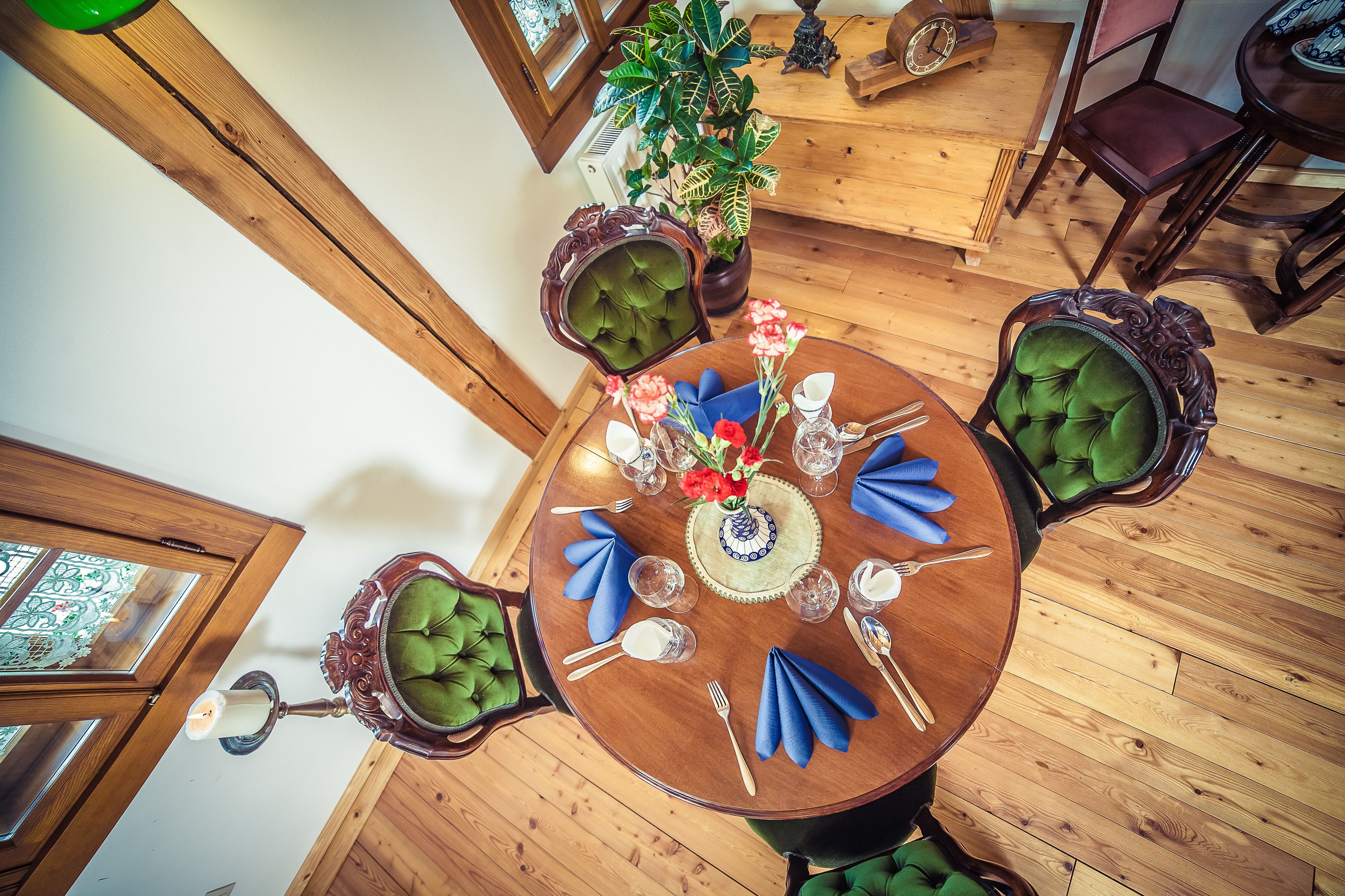 A round wooden dining table with green upholstered chairs, set with blue napkins, silverware, and a vase of flowers.