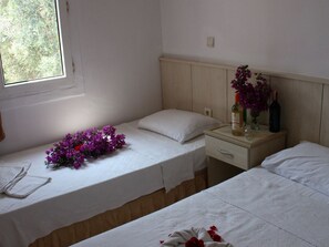 A room with two beds, a window with a view of greenery, a bedside table with a bottle and flowers, and a nightstand with a lamp. - Cennet Apart (Bodrum)