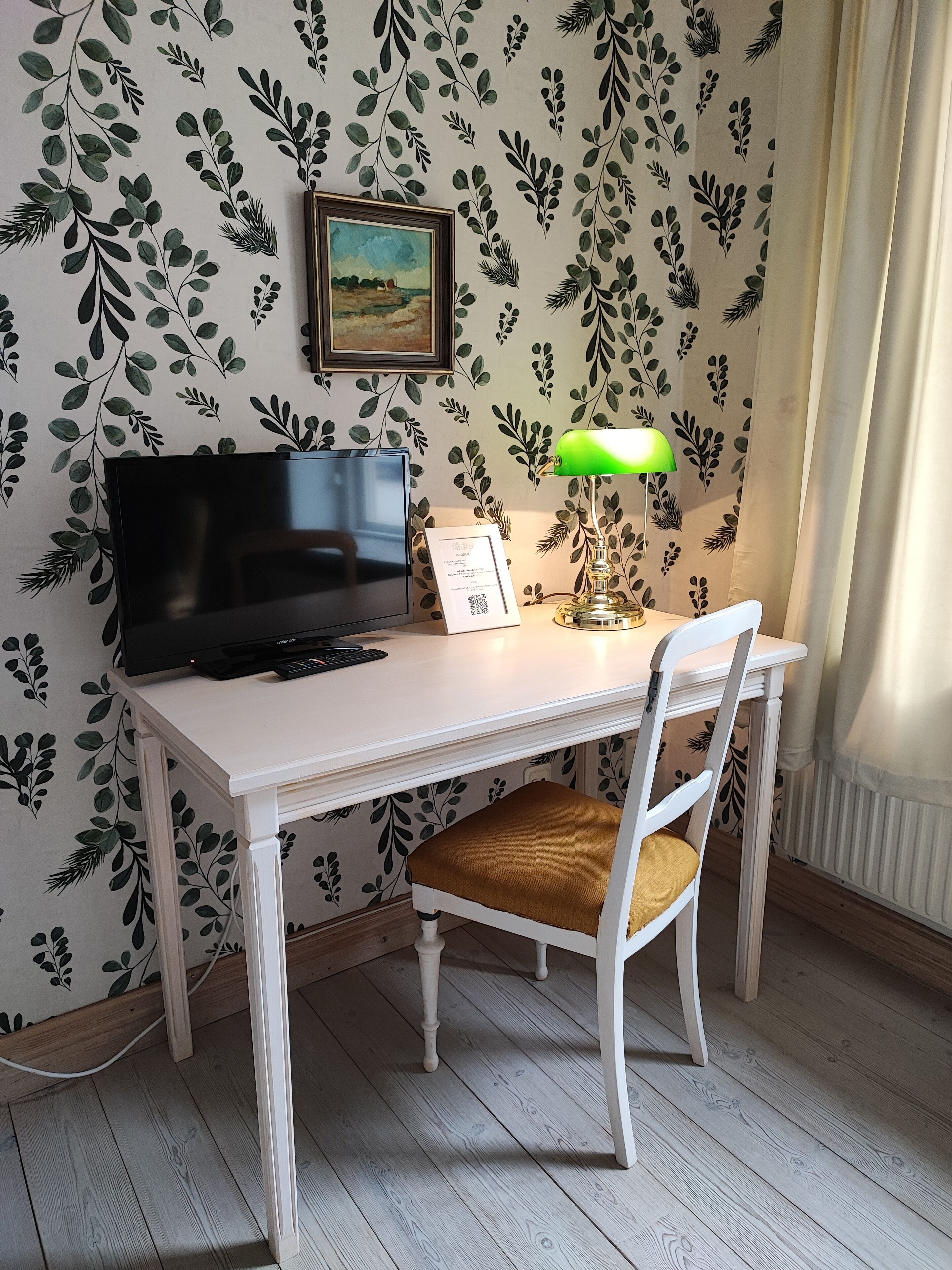Ett rum med ett vitt skrivbord, en dator, en inramad tavla och en grön bordslampa.