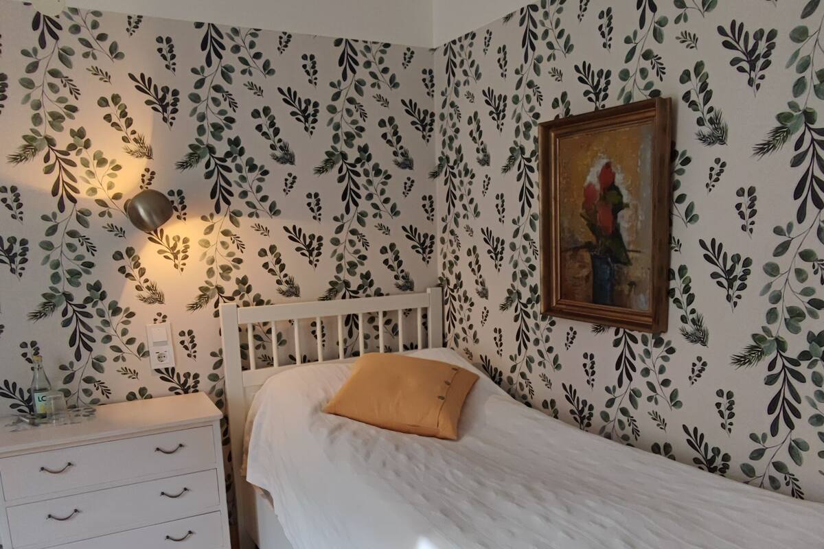 Un dormitorio con una cama blanca, una mesita de noche, un tapete verde y una pared con un diseño de hojas.