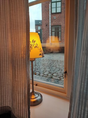 A window with a lamp on the sill, a view of a cobblestone courtyard, and a brick building in the background. - Lilla Hotellet (Lund)