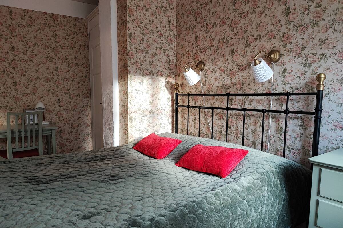 Una cama bien tendida con un edredón gris y dos almohadas rojas, una mesita de noche de madera con una lámpara, y una pared con papel tapiz floral.