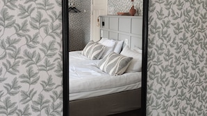 Une chambre avec un grand miroir, un lit recouvert de draps blancs et un papier peint à motifs.
