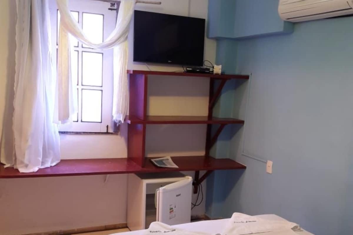 Quarto com cama, televisão, escrivaninha com computador e janela com cortinas.
