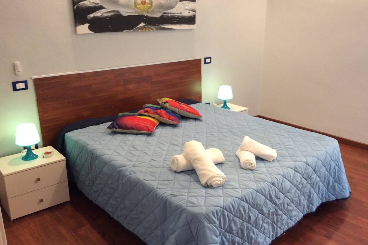 Un lit bien fait, avec une couette bleue, plusieurs oreillers et deux serviettes enroulées, le tout posé sur une tête de lit en bois.