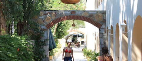 Uma rua estreita com um arco de tijolos, vegetação nas laterais e um edifício com paredes e arcos brancos.