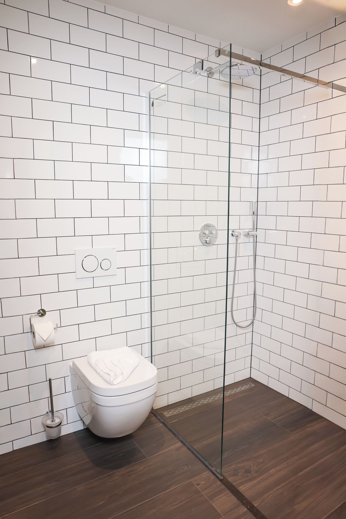 Une salle de bain moderne avec des carreaux de métro blancs, une douche en verre, des toilettes blanches et un plancher en bois foncé.
