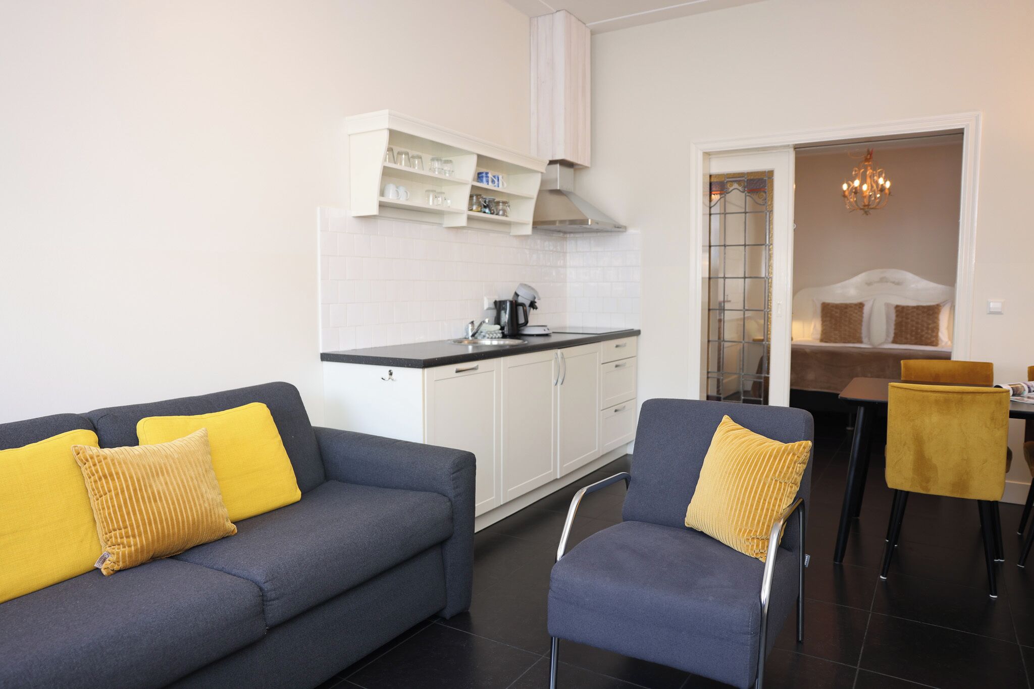 A modern living room with a grey sofa and armchair, a kitchen area with white cabinets, and a dining area with a yellow chair.