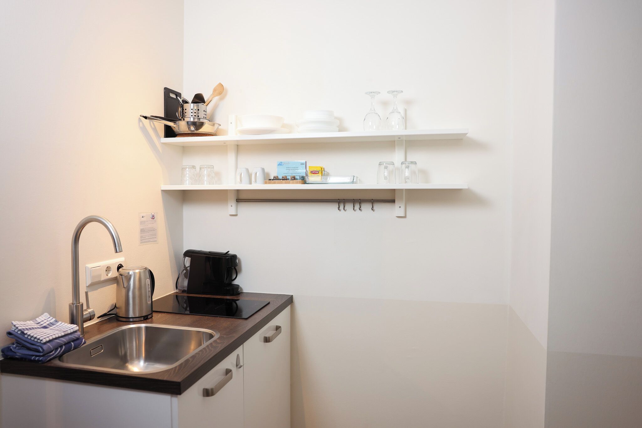 A modern kitchen with a stainless steel sink, a kettle, and a coffee maker. There are white shelves above the counter holding dishes and glasses.