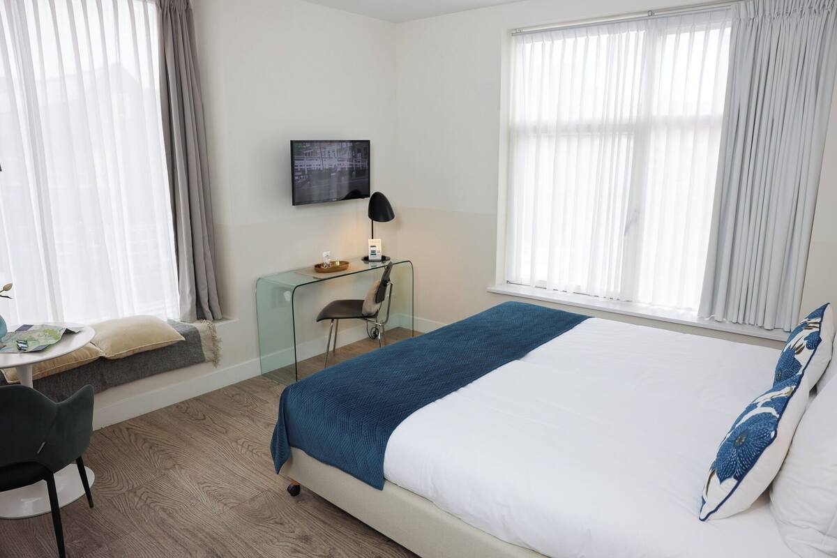 Ein Hotelzimmer mit einem Bett, einem Schreibtisch mit Stuhl, einem an der Wand befestigten Fernseher und großen Fenstern mit Vorhängen.