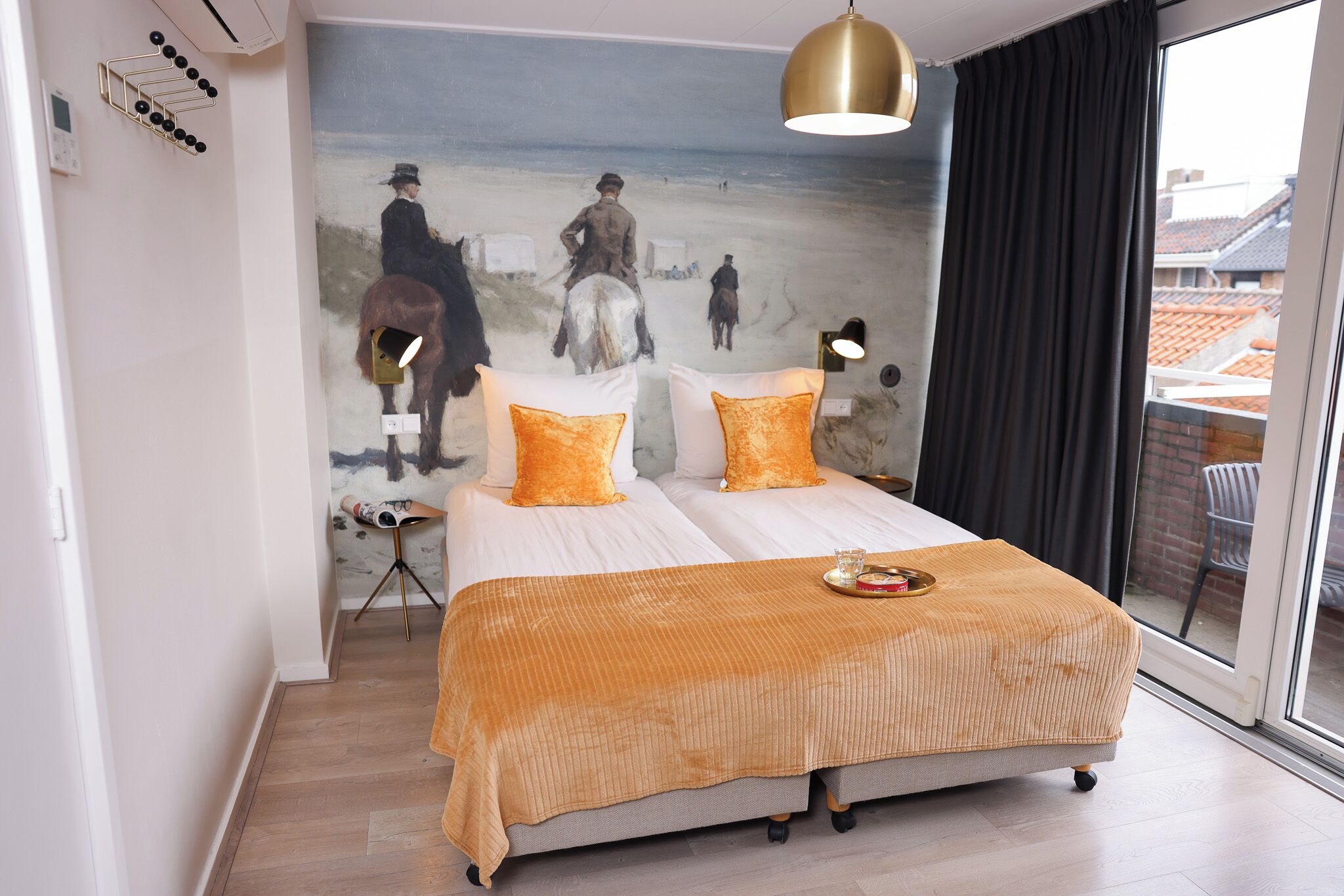 A double bed with a beige blanket and two orange pillows, a painting of a beach scene on the wall, a standing lamp, and a window with a view of buildings.