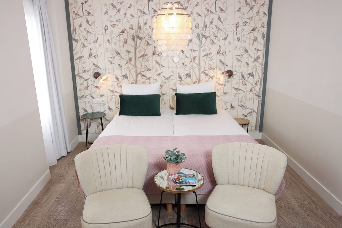 Ein Doppelbett mit zwei Kissen, ein Nachttisch mit Pflanze und zwei Sessel in einem Raum mit floralem Wandpapier und einer Pendelleuchte.