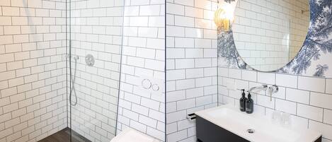 Et moderne badeværelse med hvide metrofliser, et glasbadeværelsesafskærmning, et hvidt toilet, en sort kommode med en hvid vask og et dekorativt spejl med et blomstermønster i blå farver.