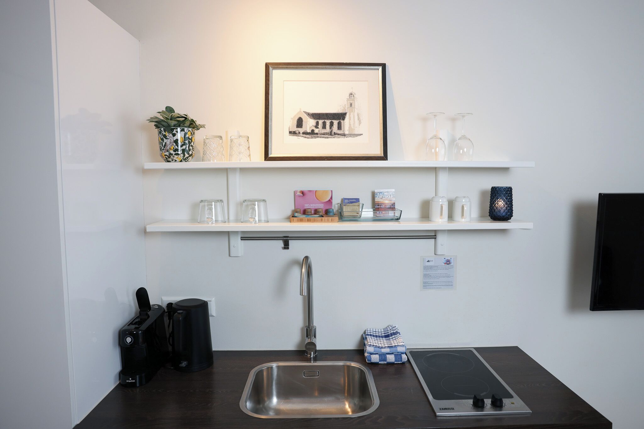A modern kitchen with a stainless steel sink, a coffee machine, and a built-in induction cooktop. There are white shelves above the sink holding various kitchen items and a framed picture.