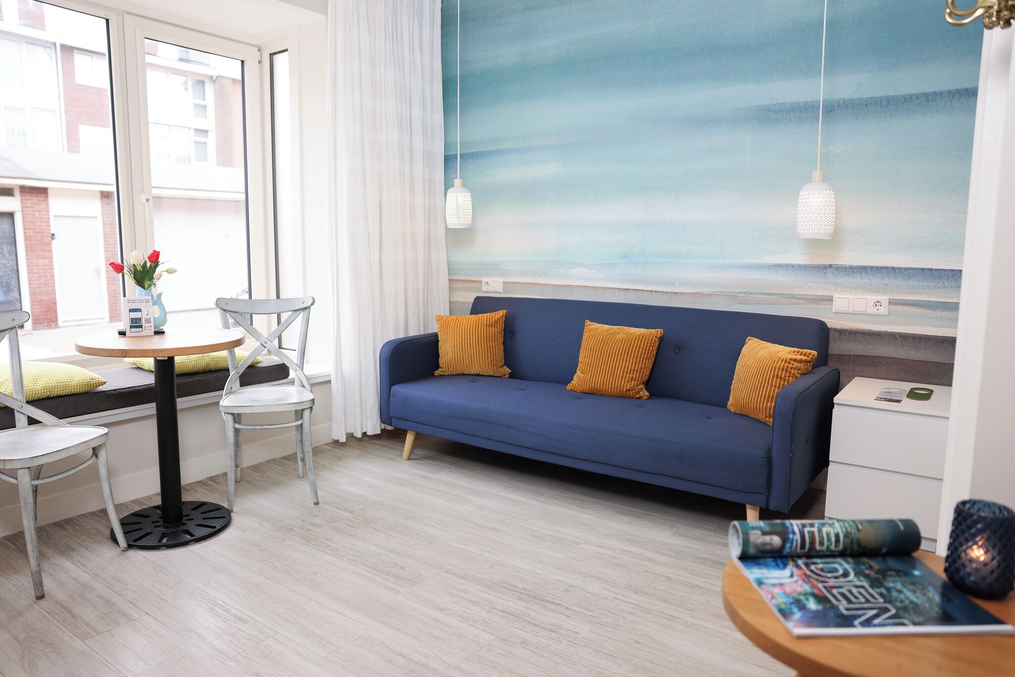 A modern living room with a blue sofa, white chairs, a round table, and a view of the city through large windows.