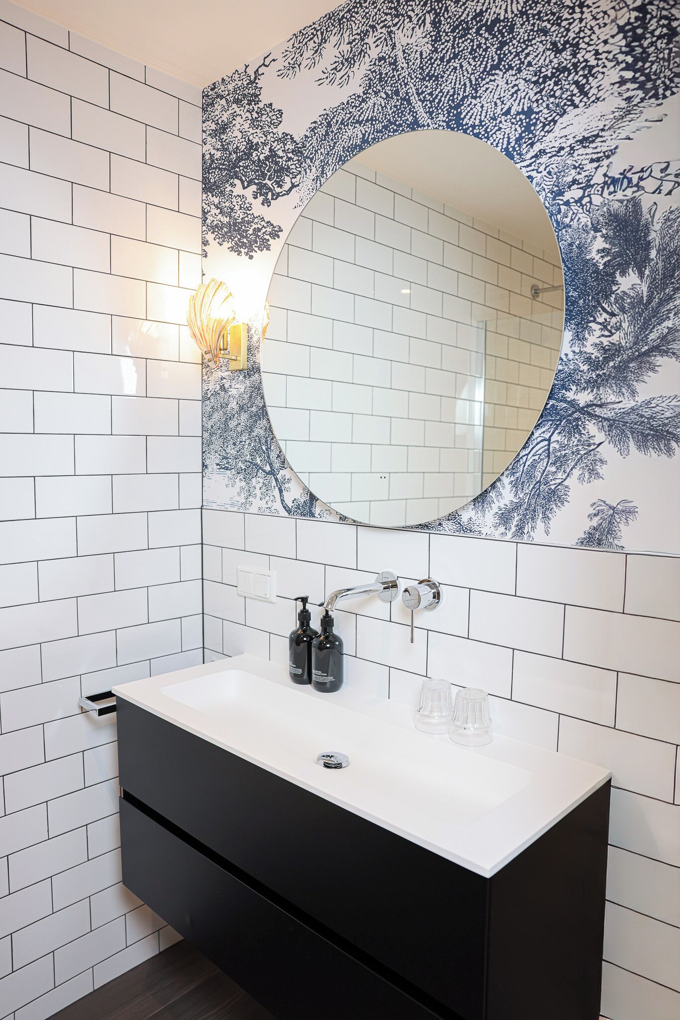 Un baño con paredes de azulejos blancos, un tocador negro, un espejo redondo y una lámpara fijada a la pared.