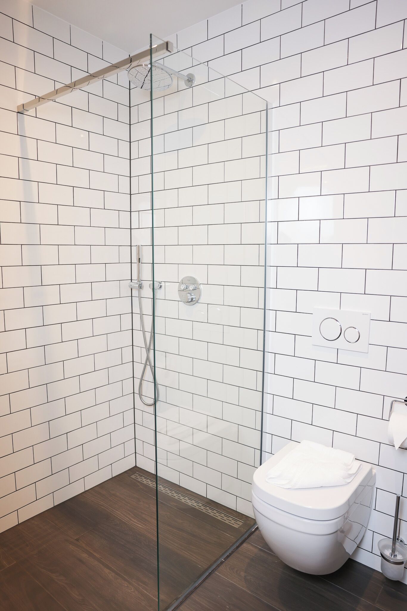 Un baño con azulejos blancos tipo metro, cerramiento de ducha de vidrio, suelo de madera y un inodoro blanco.