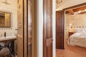 A bedroom with a wooden door, a bed with a patterned bedspread, a nightstand, and a mirror with a decorative frame. - B&B La Casa delle Rondini (Roncoferraro)