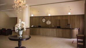 A hotel lobby with a reception desk, two wall clocks, a chandelier, and a round table with a vase of flowers. - Hotel Premier (São Luís)