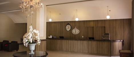 A hotel lobby with a reception desk, two wall clocks, a chandelier, and a round table with a vase of flowers.