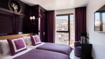 Habitación de hotel con dos camas, un ventanal grande, vistas a edificios y un televisor sobre un mueble.