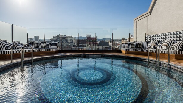 A rooftop pool with clear blue water, surrounded by black and white striped lounge chairs and a cityscape view in the background. - Room Mate Anna, Barcelona (Barcelona)