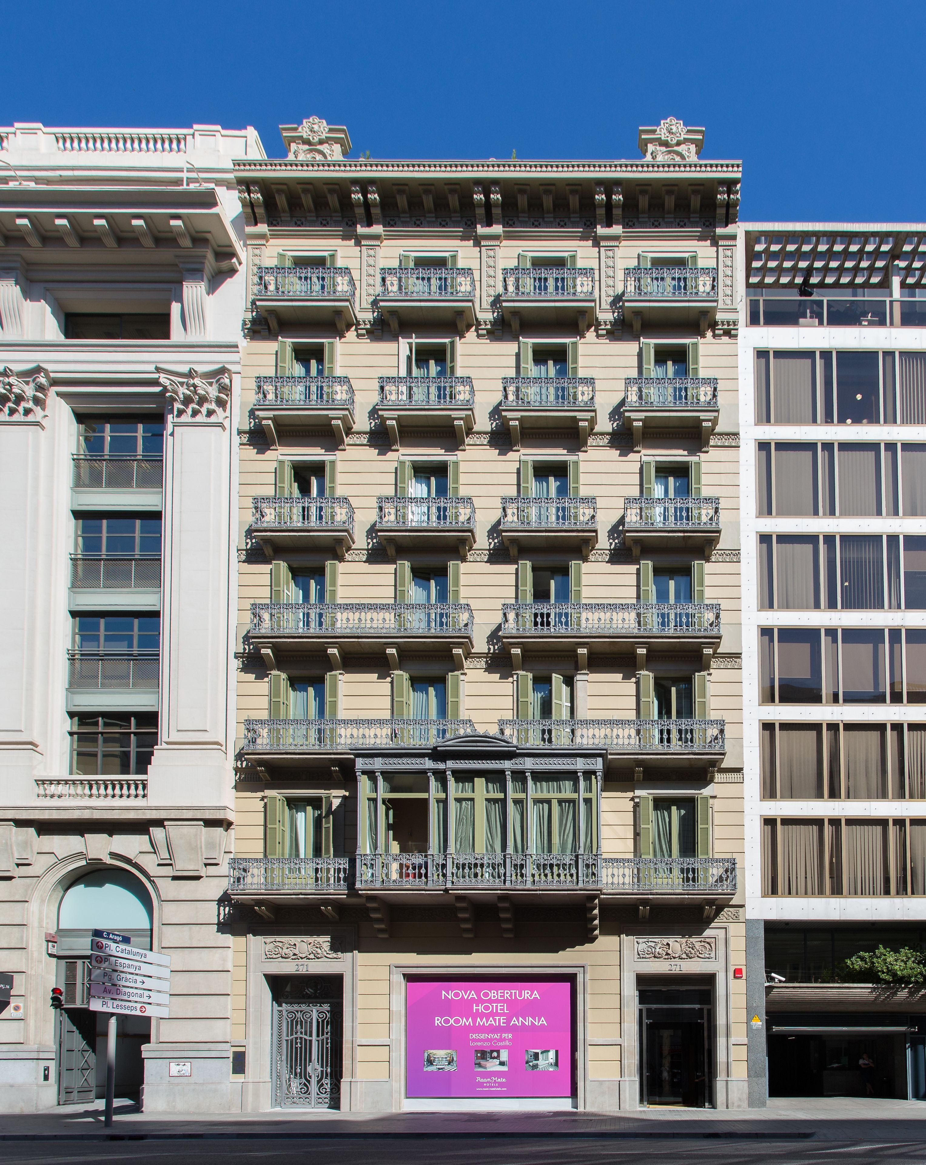 Un hotel de varios pisos con balcones, un letrero llamativo que dice "NOVA OBTURADA HOTEL ROOM MATE ANNA" y una señal de calle que indica la dirección a puntos de referencia cercanos.