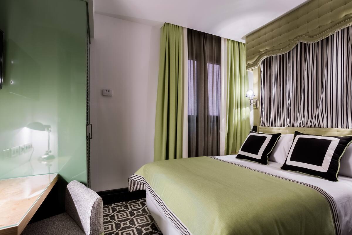 A hotel room with a bed, a desk, and a chair. There is a window with curtains and a wall-mounted lamp.