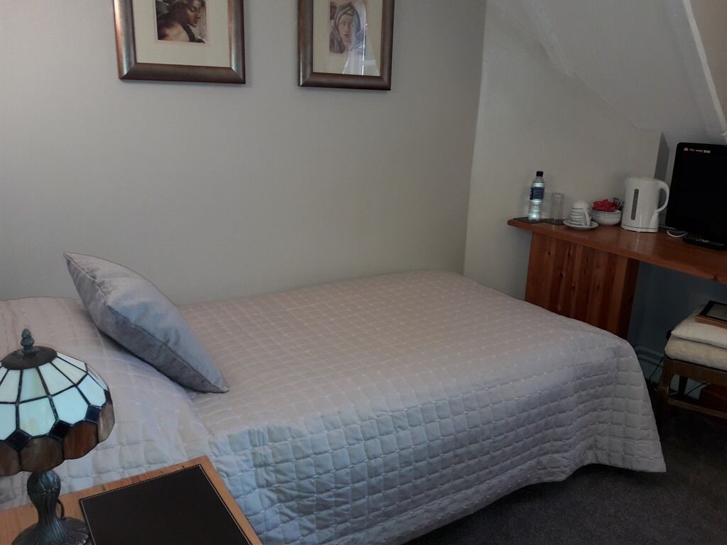 Un dormitorio con una cama, una mesita de noche con lámpara, un pequeño escritorio con computadora y dos cuadros en la pared.