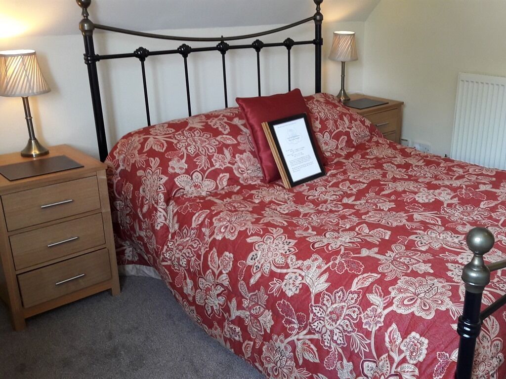 A neatly made bed with a floral-patterned bedspread and a single red pillow. There is a bedside table with a lamp and a framed certificate or notice. The room has a simple, clean design with a radiator on the wall.