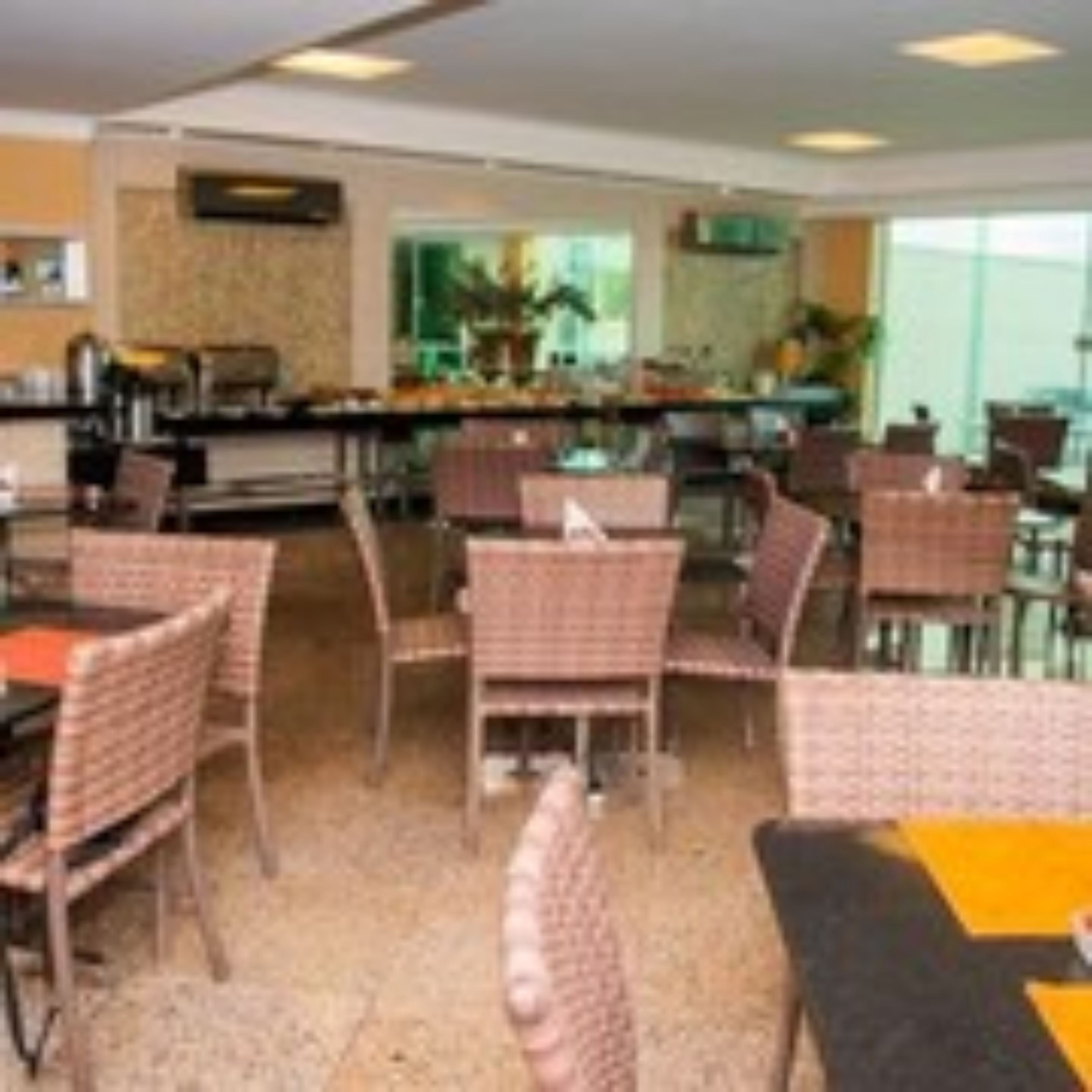 Comedor de hotel con sillas y mesas de mimbre, zona de bar y vistas a la cocina a través de un muro de cristal.