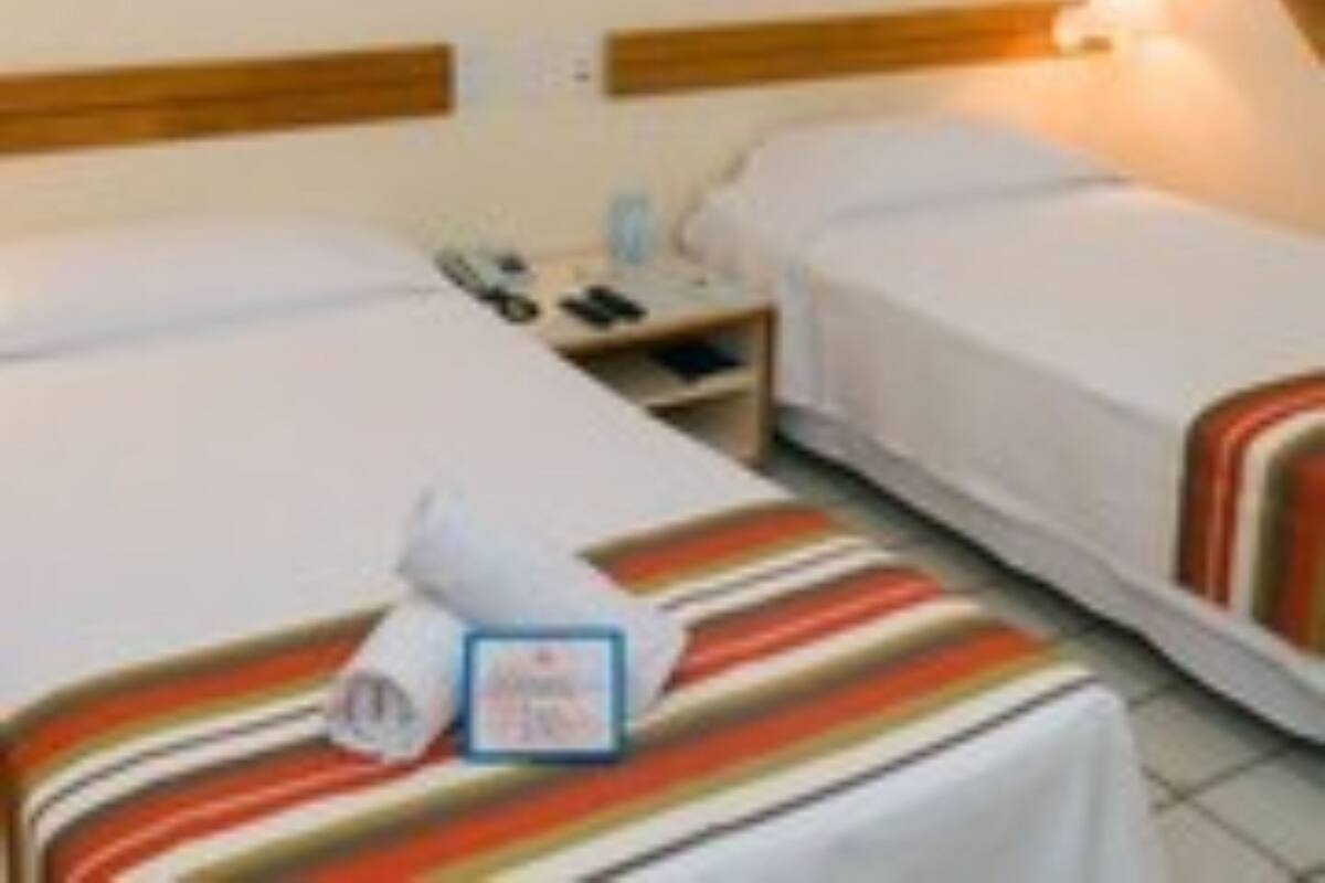 Quarto de hotel com duas camas de solteiro, mesa de cabeceira, abajur e uma pequena mesa.