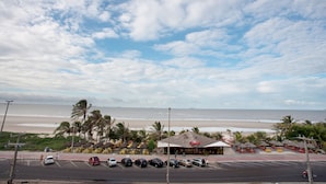 A beachside parking lot with cars, a restaurant with outdoor seating, and a view of the ocean. - Calhau Praia Hotel (São Luís)