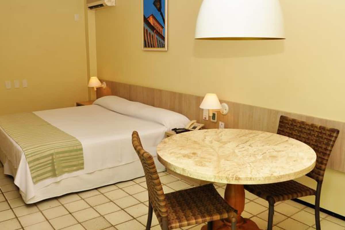 Quarto de hotel com uma cama, uma mesa redonda, duas cadeiras, um abajur e um quadro na parede.