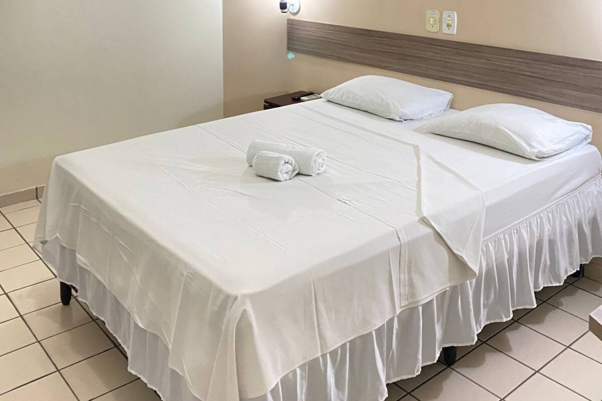 A hotel room with a bed, a nightstand, a wall-mounted air conditioner, and a wall socket.