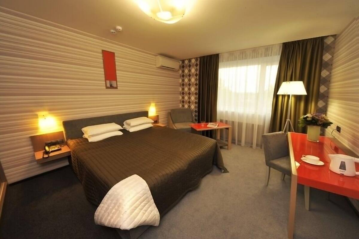 Ein Hotelzimmer mit einem großen Bett, einem Schreibtisch mit Stuhl, einer Lampe und einem kleinen Tisch mit Pflanze.