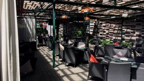 An outdoor seating area with wicker chairs and tables, a patterned wall, and a green metal structure overhead. - Villa Four Rooms (Kharkiv)