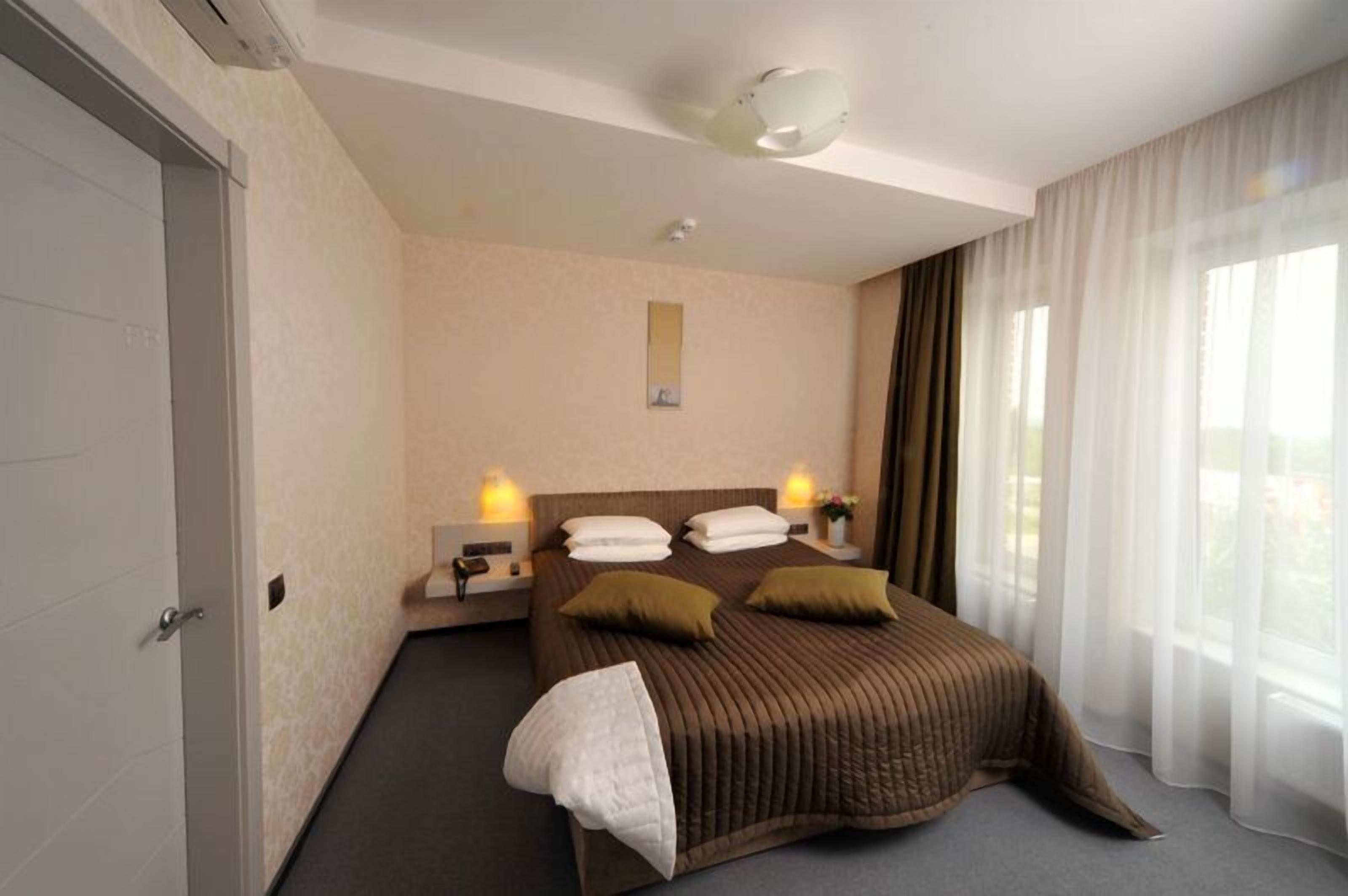 Habitación de hotel con una cama grande, dos mesitas de noche con lámparas, una mesita pequeña con un teléfono, una ventana con cortinas traslúcidas y una luz en el techo.