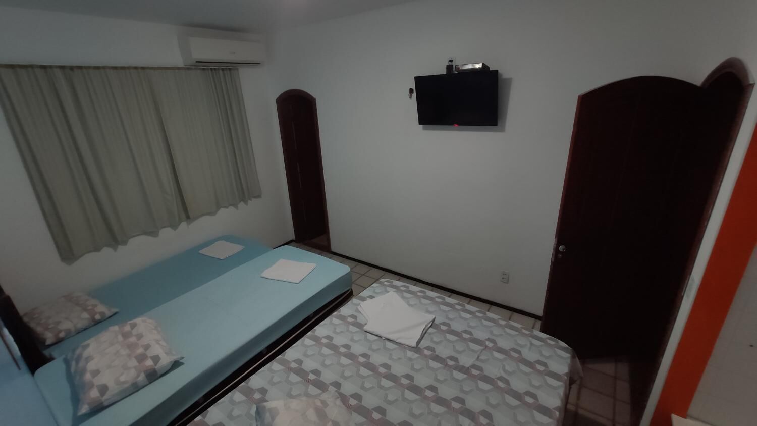 Una habitación con dos camas, una con colchón azul y la otra con un estampado. Hay un televisor montado en la pared, una ventana con cortinas y una puerta.