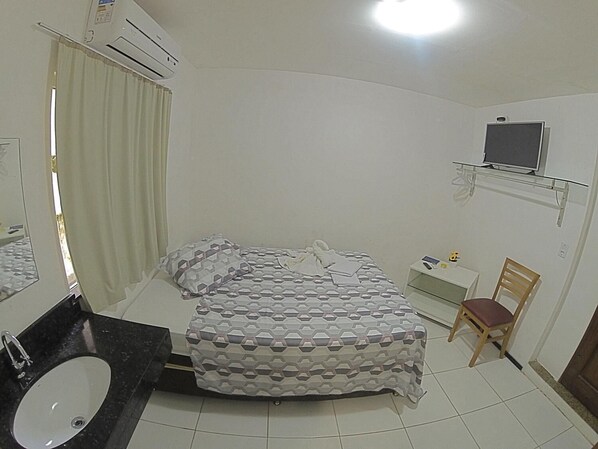 Une chambre d’hôtel avec un lit, un bureau, une chaise, un téléviseur posé sur une étagère et une salle de bain avec un lavabo.