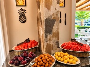 Un bufé con diversas frutas y frutos secos, una decoración de piña y vistas al área de mesas al aire libre.