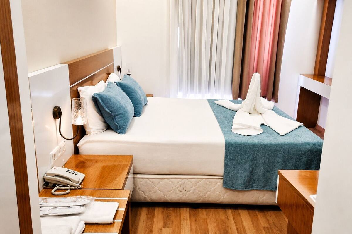 Een hotelkamer met een bed, nachtkastje met telefoon, lamp en raam met gordijnen.