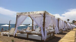 A beachside resort with rows of white canopy beds and lounge chairs, a wooden walkway, and umbrellas.